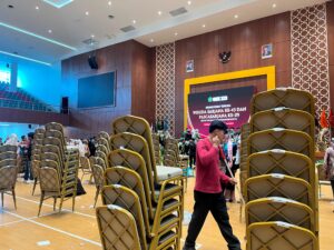 Seorang petugas kebersihan sedang membereskan dan menumpuk kursi di dalam aula usai prosesi wisuda selesai, di saat para wisudawan dan keluarga masih sibuk berswafoto. Sabtu,08/08/2026.